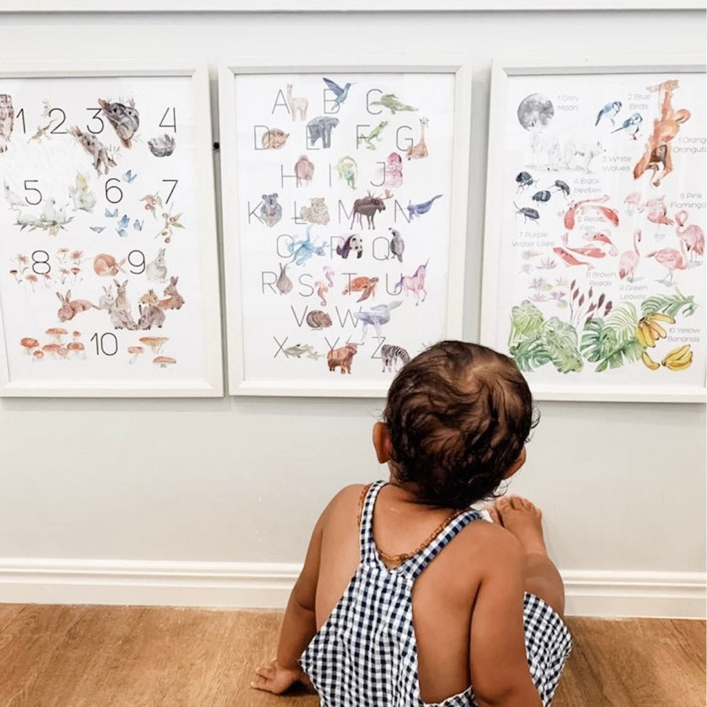 baby sitting down on a hardwood floor looking at three framed illustrated wall art prints in their playroom
